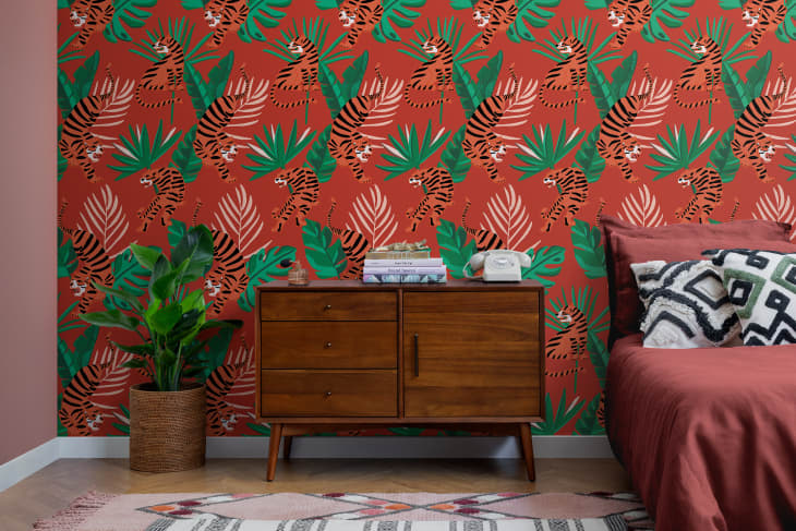 Bedroom with red and green tropical wallpaper, wooden dresser, potted plant, and red bedding with patterned pillows.