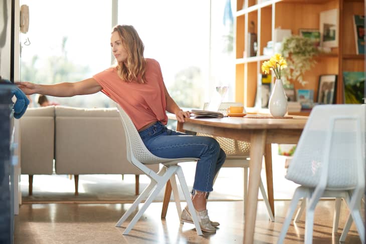 Woman in a coral shirt sitting at a wooden dining table with a vase of yellow flowers, reaching towards a shelf.