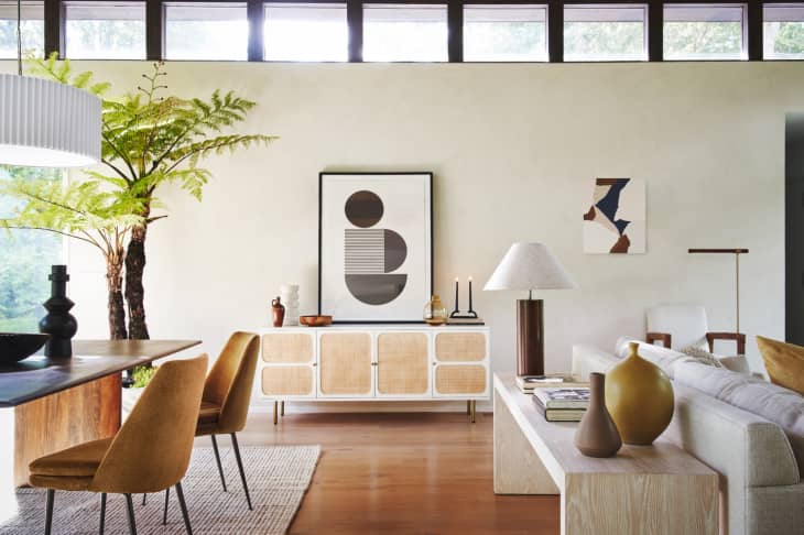 Modern living room with abstract art, wooden sideboard, mustard chairs, and a large potted fern.