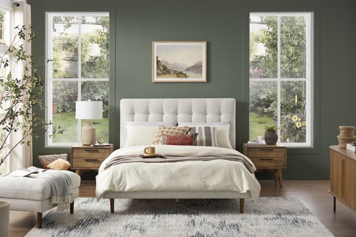 Bedroom with green walls, tufted headboard, wooden nightstands, and large windows overlooking a garden.
