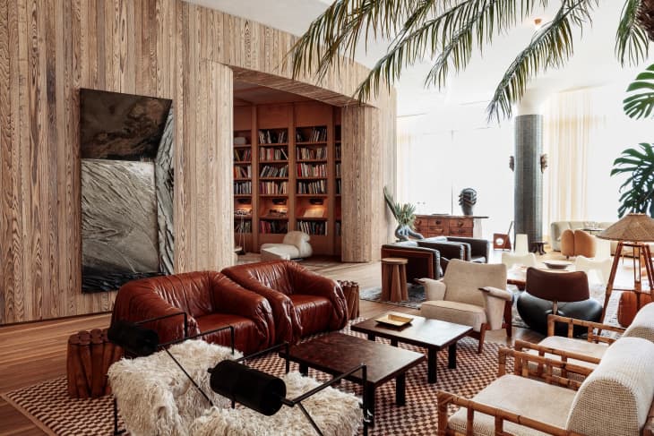 Spacious living room with leather sofas, wooden walls, large bookshelf, and tropical plants.