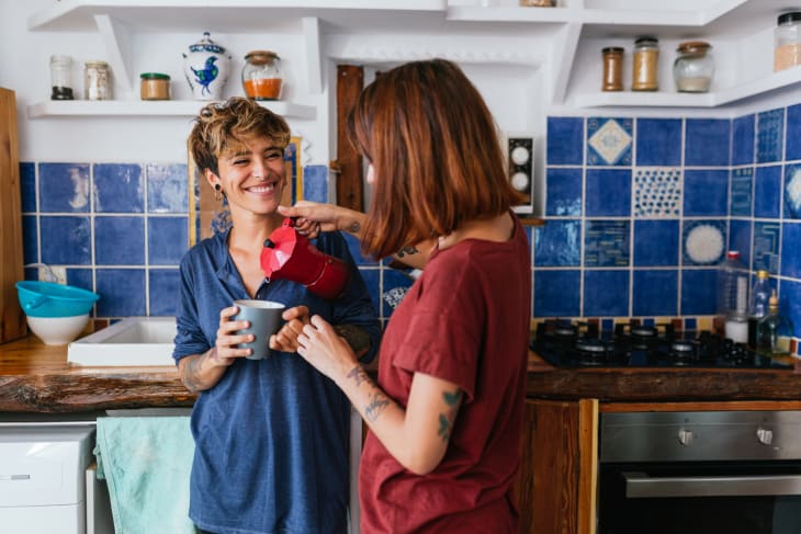 Two people in a kitchen with blue tiles, one pouring coffee from a red pot into a mug.