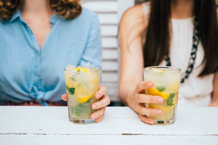 Two women holding glasses of lemonade with mint and lemon slices at a white table.