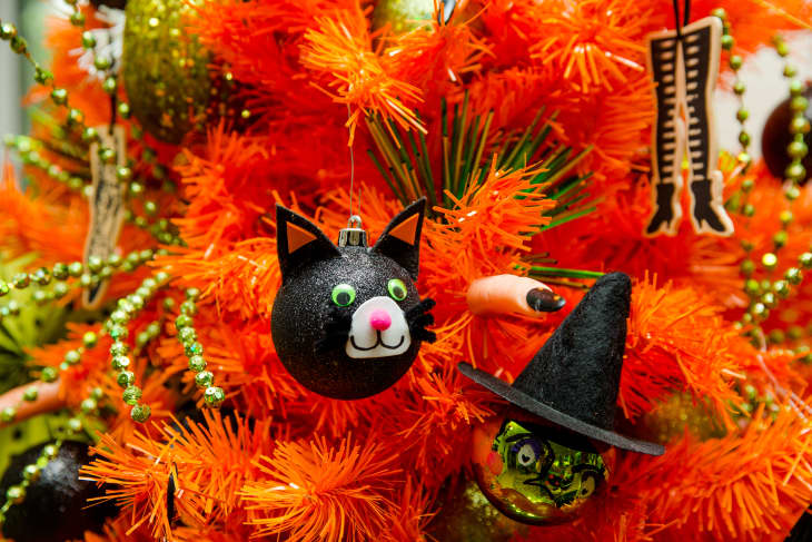 Halloween-themed ornaments on an orange Christmas tree, featuring a black cat, witch face, and striped legs.