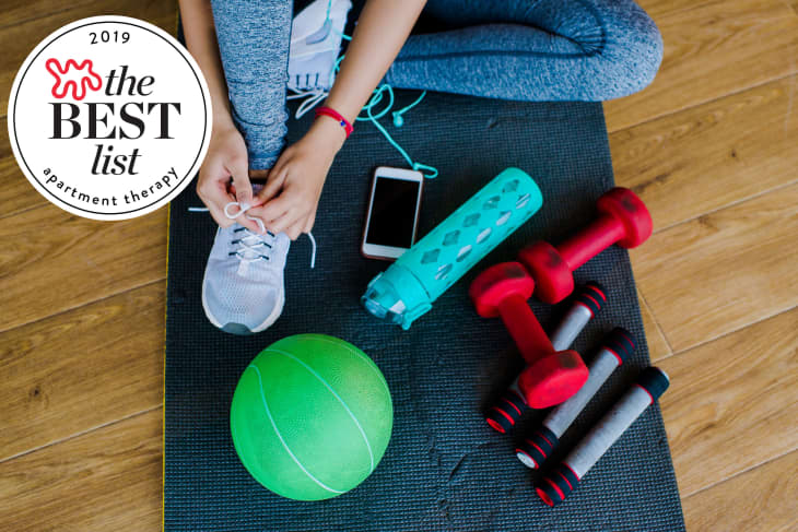 Person tying sneakers on a yoga mat with dumbbells, a green ball, a water bottle, and a smartphone nearby.