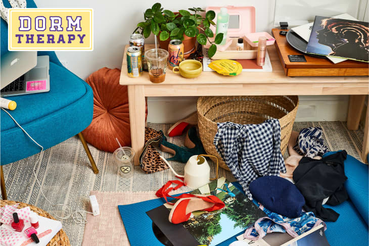 Cluttered dorm room with a blue chair, wooden table, plants, drinks, vinyl records, and scattered clothes on the floor.