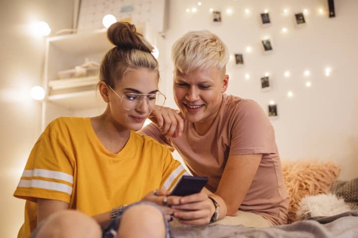 Two people sitting on a bed, smiling at a smartphone, with string lights and photos on the wall.