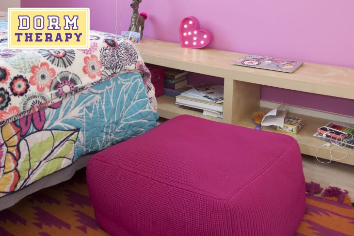 Colorful dorm room with floral bedspread, pink ottoman, heart-shaped light, and wooden shelf with books and decor.