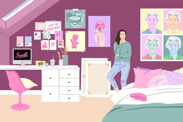 Woman in a purple room with art on walls, standing by a bed and desk with a computer and pink chair.