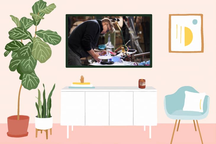 Living room with a white sideboard, potted plants, a teal chair, and a wall-mounted TV showing a person working.