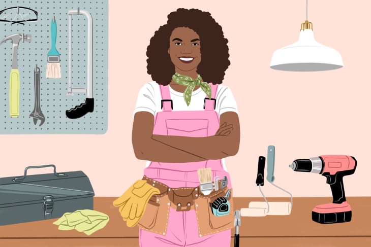 Woman in pink overalls with tool belt, standing in workshop with tools on pegboard and table, under a hanging lamp.