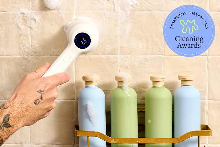 A hand holding a white cleaning device against a tiled bathroom shower wall with soap suds, next to a gold rack holding colorful bottles.