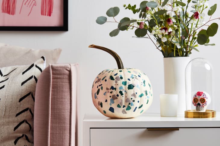 Speckled pumpkin on a white table with a vase of flowers, candle, and decorative skull under a glass dome.