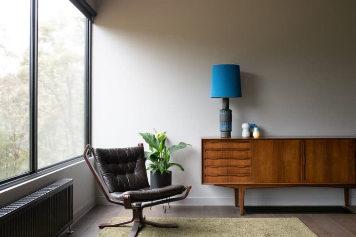 Mid-century modern room with a leather chair, wooden sideboard, blue lamp, and potted plant by a large window.