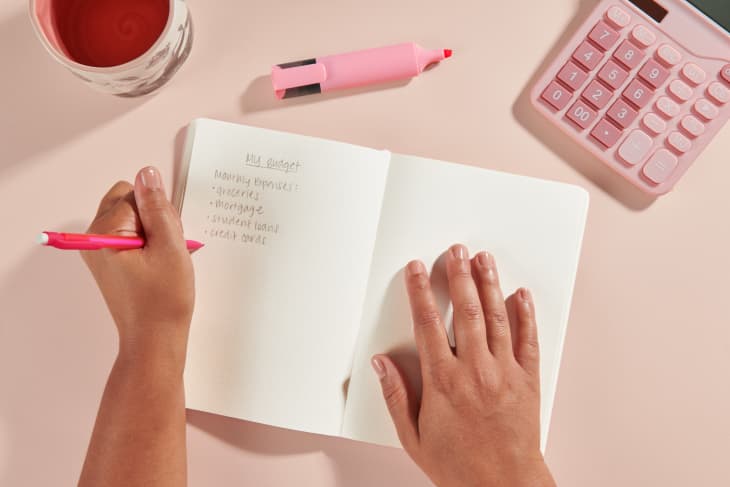 Writing budget notes into a notebook with a pink calculator, mug, highlighter on a pink background