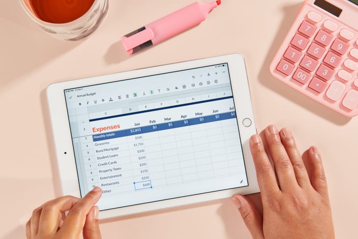 entering expenses into a budget spreadsheet on an ipad with a pink background, calculator, and mug
