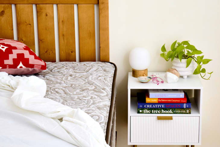 a mattress with sheets coming off next to a nightsand with a lamp, plant, books, and glasses.