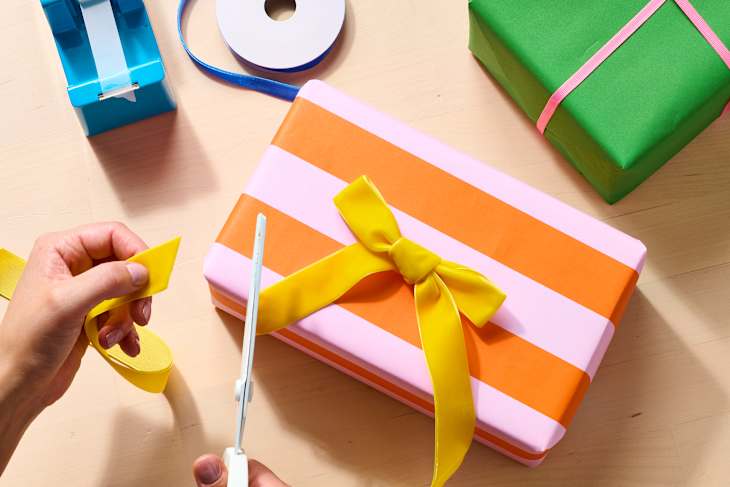 A person cutting a yellow ribbon while wrapping a pink and orange striped gift box, with green and blue gifts nearby.