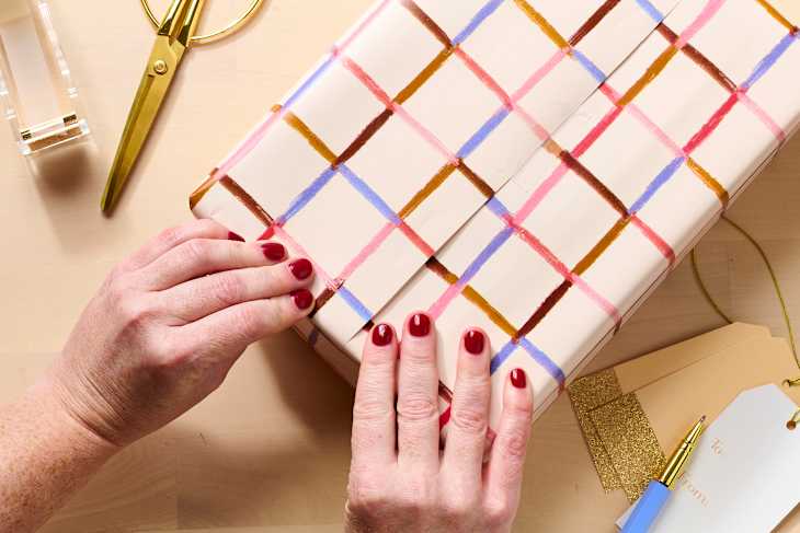 Hands with red nails wrapping a gift in patterned paper, surrounded by scissors and gift tags.