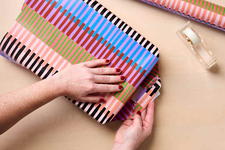 Hands with red nail polish wrapping a gift in colorful striped paper, with a tape dispenser nearby.