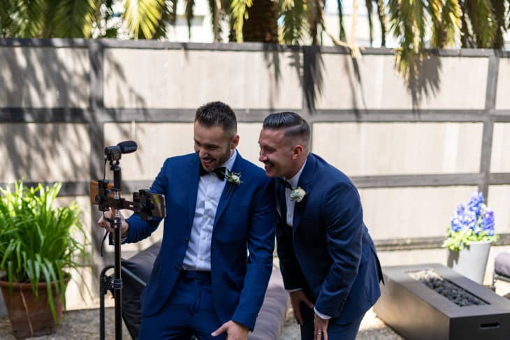 Two men in blue suits smiling at a phone on a tripod in a garden setting with plants and a wooden fence.