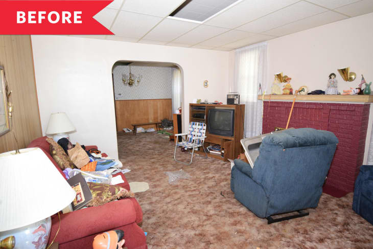 Cluttered living room with red sofa, blue recliner, old TV, and brick fireplace. Archway leads to dining area with chandelier.