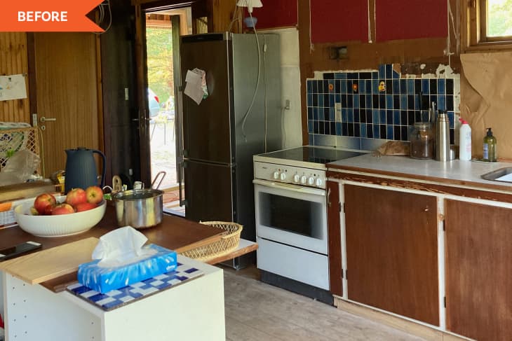"before" photo of a kitchen under renovation. There is a fridge, a stove, some a blue tile backsplash. The cabinets are all dark wood laminate. There are cords hanging from the ceiling. To the left of the photo there is bowl of apples, a tea kettle, a saucepan, and a box of facial tissues. Orange “BEFORE” flag is in upper left corner