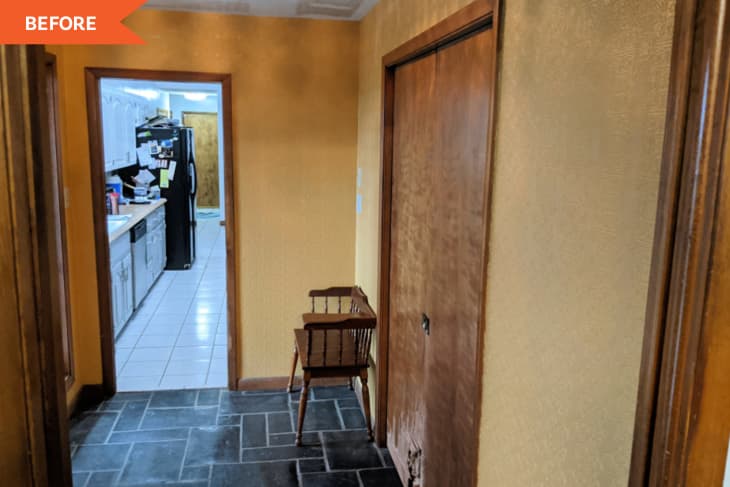 Hallway with yellow walls, wooden doors, a small wooden chair, and view into a kitchen with white cabinets and black fridge.