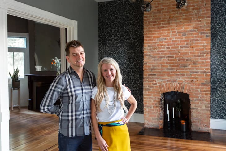 Dana and her friend are standing together in front of a brick fireplace in a living room, smiling.