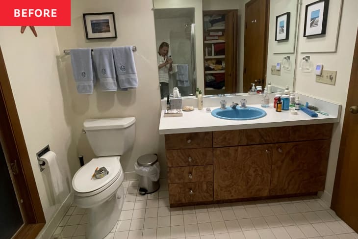 Bathroom before renovation. dated faux wood cabinets, white counter, blue sink, off white walls, white tile floor