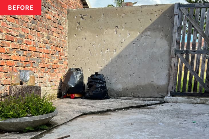 Patio before decorating/renovation. One red brick wall, another plain gray wall, a couple plants, 2 trash bags, no furniture