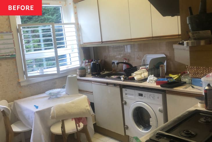 Dated kitchen with small cloth covered dining table near window and neutral colored cabinets.