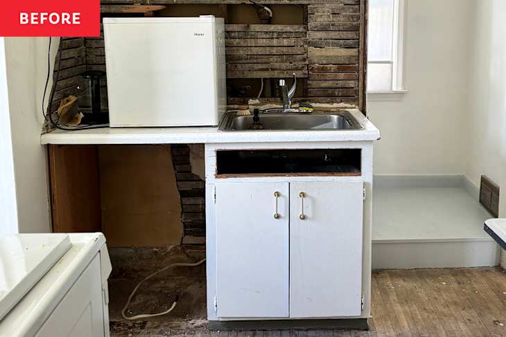 A small kitchen area with a white mini fridge, sink, and open cabinet space, featuring exposed wooden walls and unfinished flooring.
