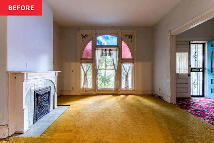 Empty living room with yellow carpet, ornate fireplace, large windows with colorful glass, and a blue door.