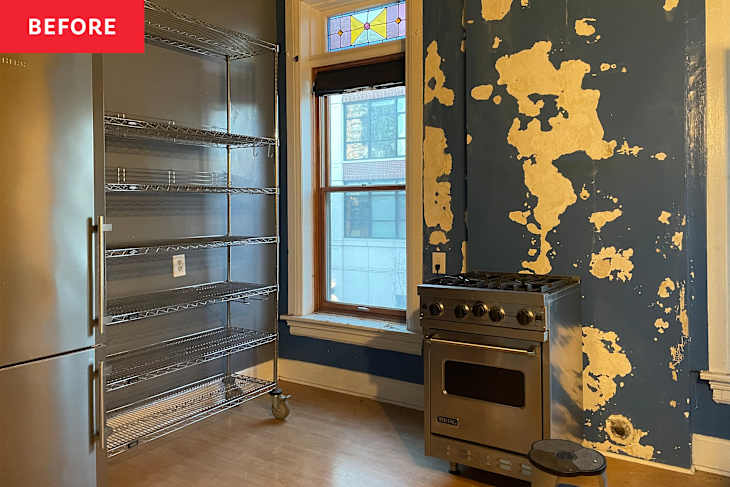 Kitchen space with a stainless steel stove, metal shelving unit, and a window with stained glass, featuring peeling blue paint on the wall.