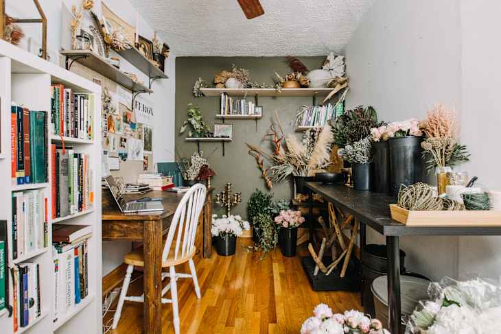 Home office with wooden desk, white chair, bookshelves, and a table filled with dried flowers and plants.