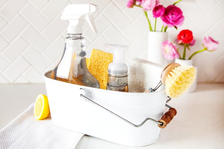 A cleaning caddy filled with spray bottles, sponges, and other cleaning supplies is pictured in front of two white vases