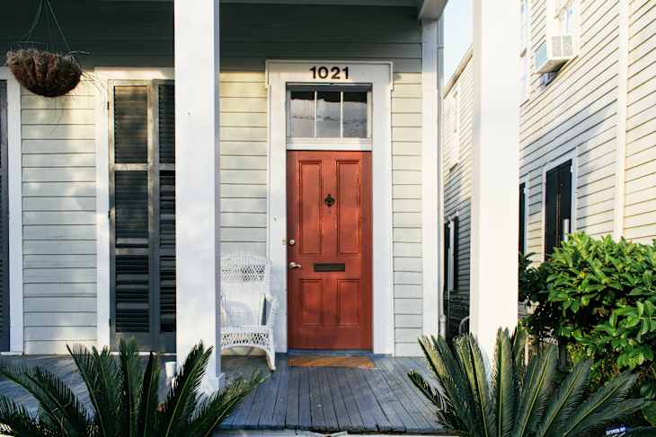 Red front door with number 1021, white wicker chair, and potted plants on a porch.