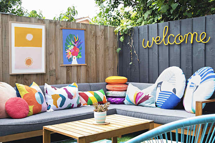 Outdoor patio with gray sectional, colorful pillows, wooden table, and "welcome" sign on fence. Two vibrant wall art pieces.
