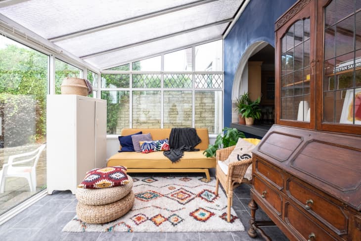 Sunroom with yellow sofa, colorful cushions, wicker chair, wooden cabinet, and patterned rug under a glass roof.