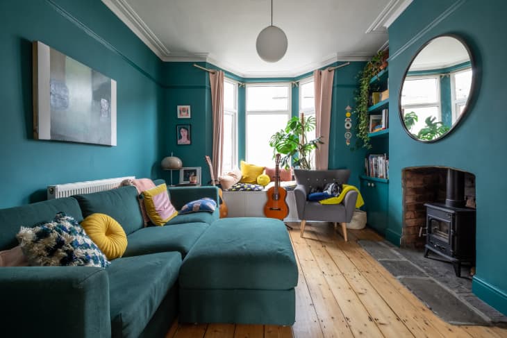 Teal living room with sectional sofa, yellow pillows, guitar, armchair, plants, and a round mirror above a fireplace.
