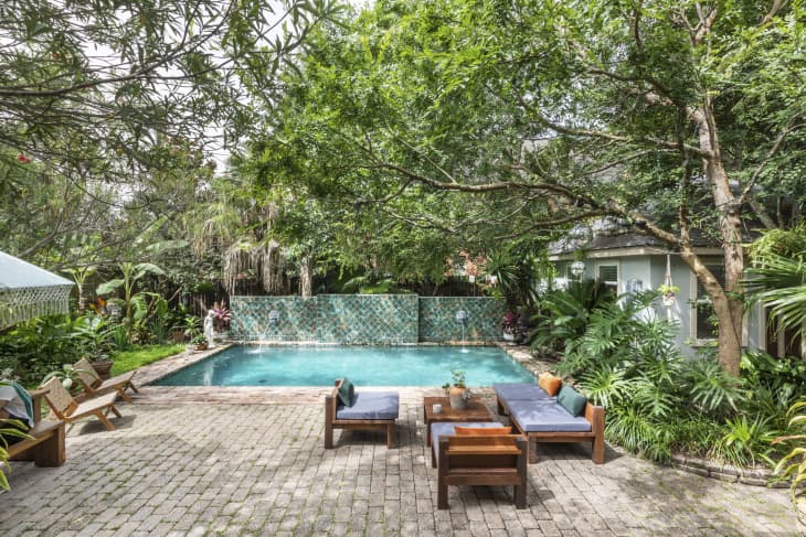 Backyard with a turquoise pool, surrounded by lush greenery, brick patio, and wooden outdoor furniture with cushions.
