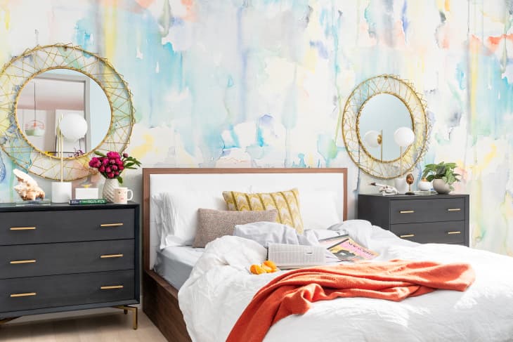 Bedroom with abstract watercolor wall, two black dressers, round mirrors, and a bed with orange throw and decorative pillows.