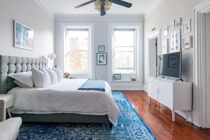 Bright bedroom with a gray tufted bed, blue rug, white dresser, and wall art, featuring large windows and a ceiling fan.