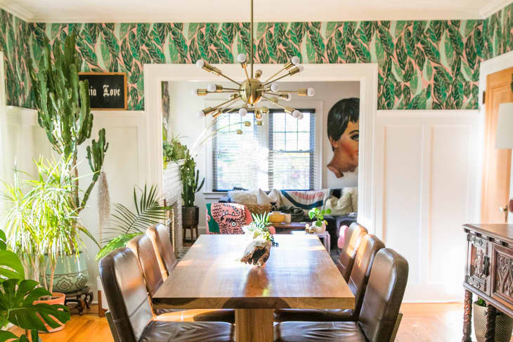 Dining room with a wooden table, leather chairs, green plants, and tropical wallpaper, leading to a cozy living area.