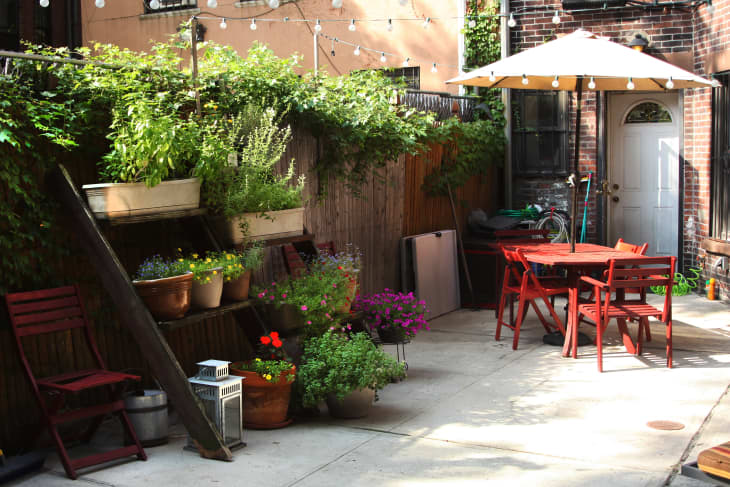 Brick patio with red table and chairs, potted plants, string lights, and a large umbrella.