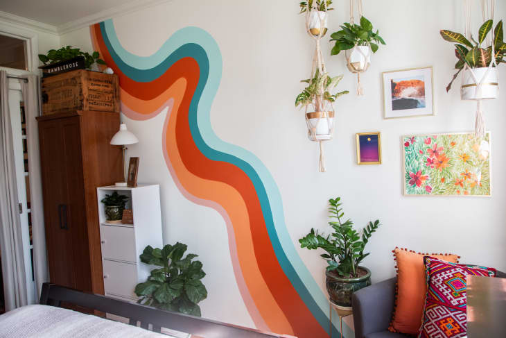 Colorful room with wavy wall mural, hanging plants, wooden cabinet, and vibrant cushions on a gray sofa.