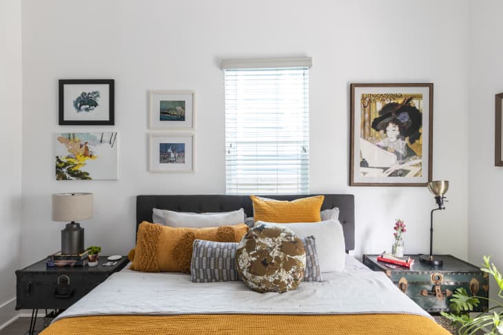 Bedroom with a dark headboard, yellow and white pillows, eclectic wall art, and vintage side tables with decor items.