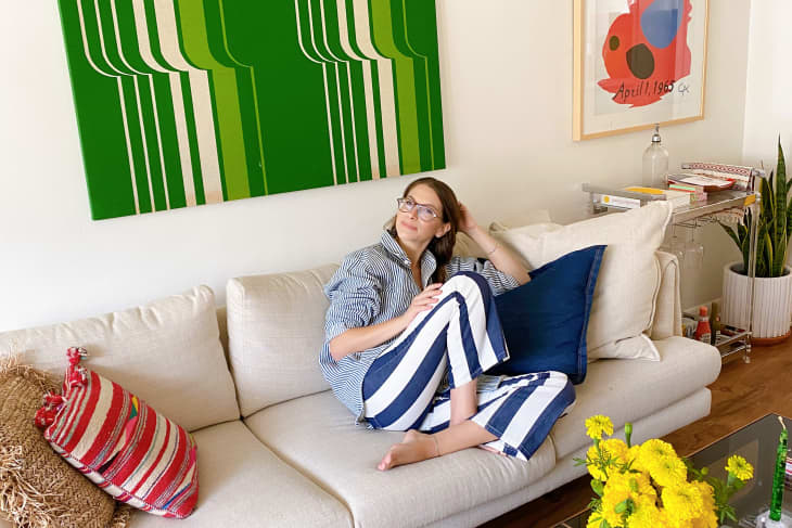 穿着蓝白条纹短裤的女人坐在米色沙发上，墙上挂着一幅20世纪70年代的绿色艺术品
