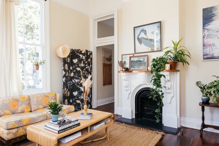 Living room with floral sofa, rattan coffee table, plants, and art above a white fireplace.